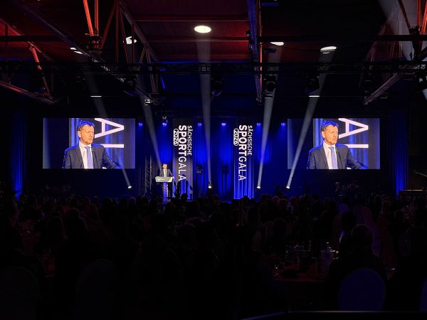 Speaker at a podium on a dark conference stage, flanked by blue vertical banners and two large screens showing him to the audience.