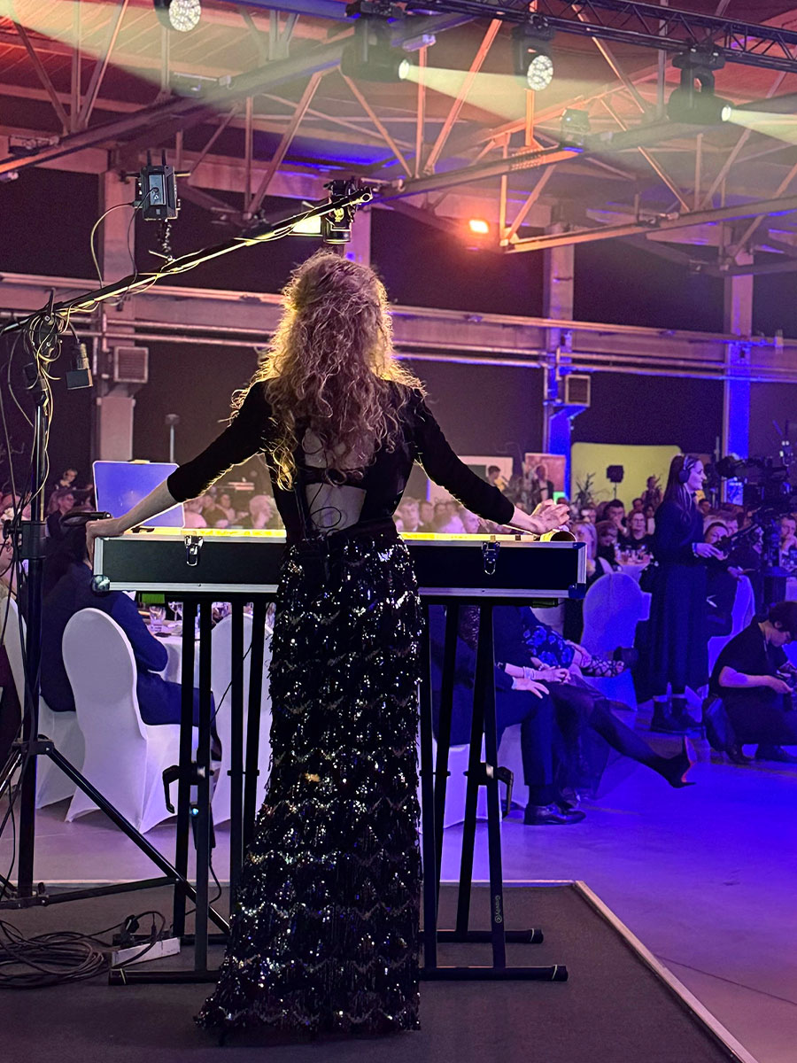 Performer with long curly hair in a glittery black gown at a keyboard on stage, colorful lights, audience seated in a banquet hall backdrop.